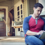 A man sitting relaxing in a quiet corner of a porch, using a digital tablet.